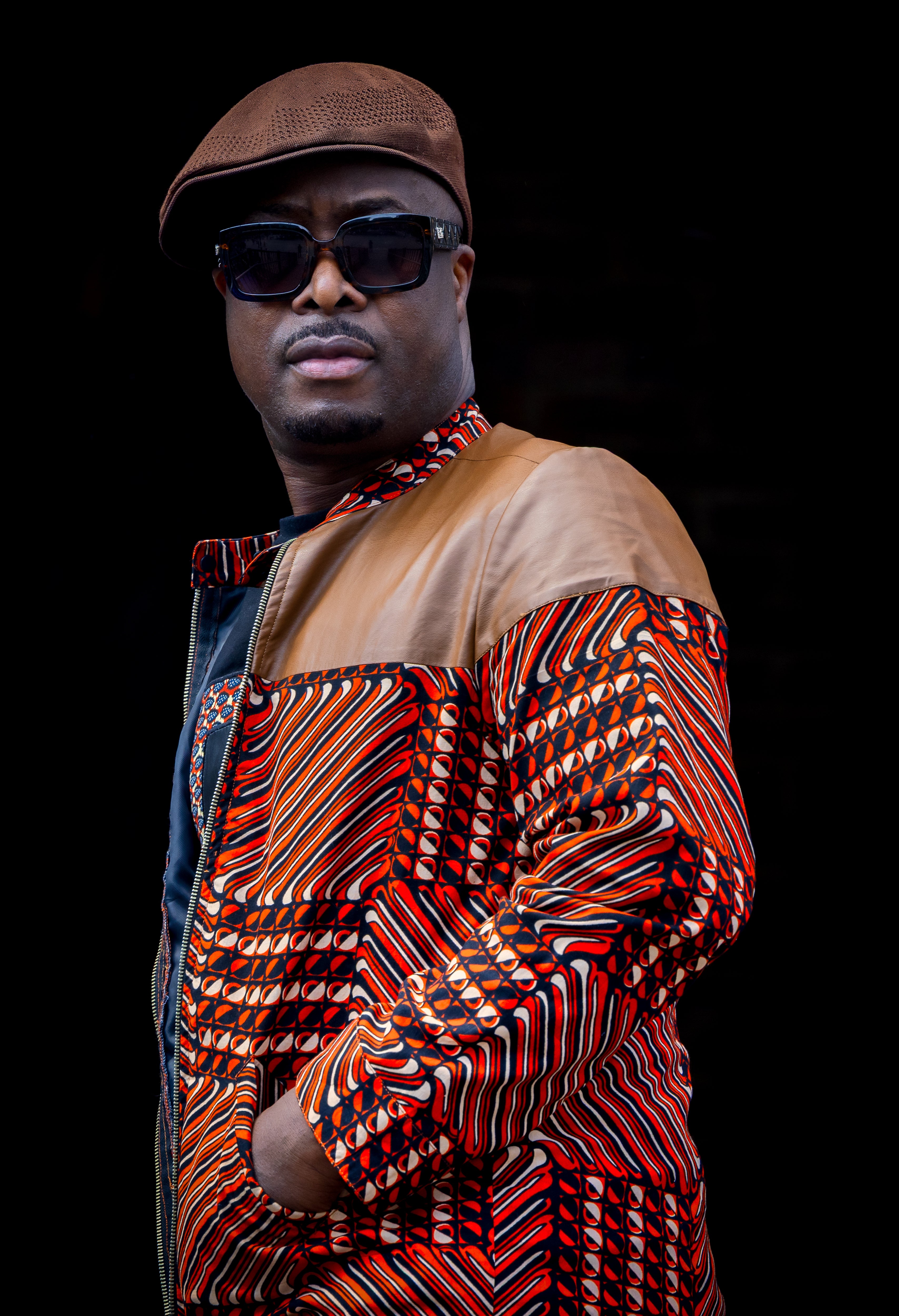 Man wearing a patterned African print jacket outfit and sunglasses against a black background